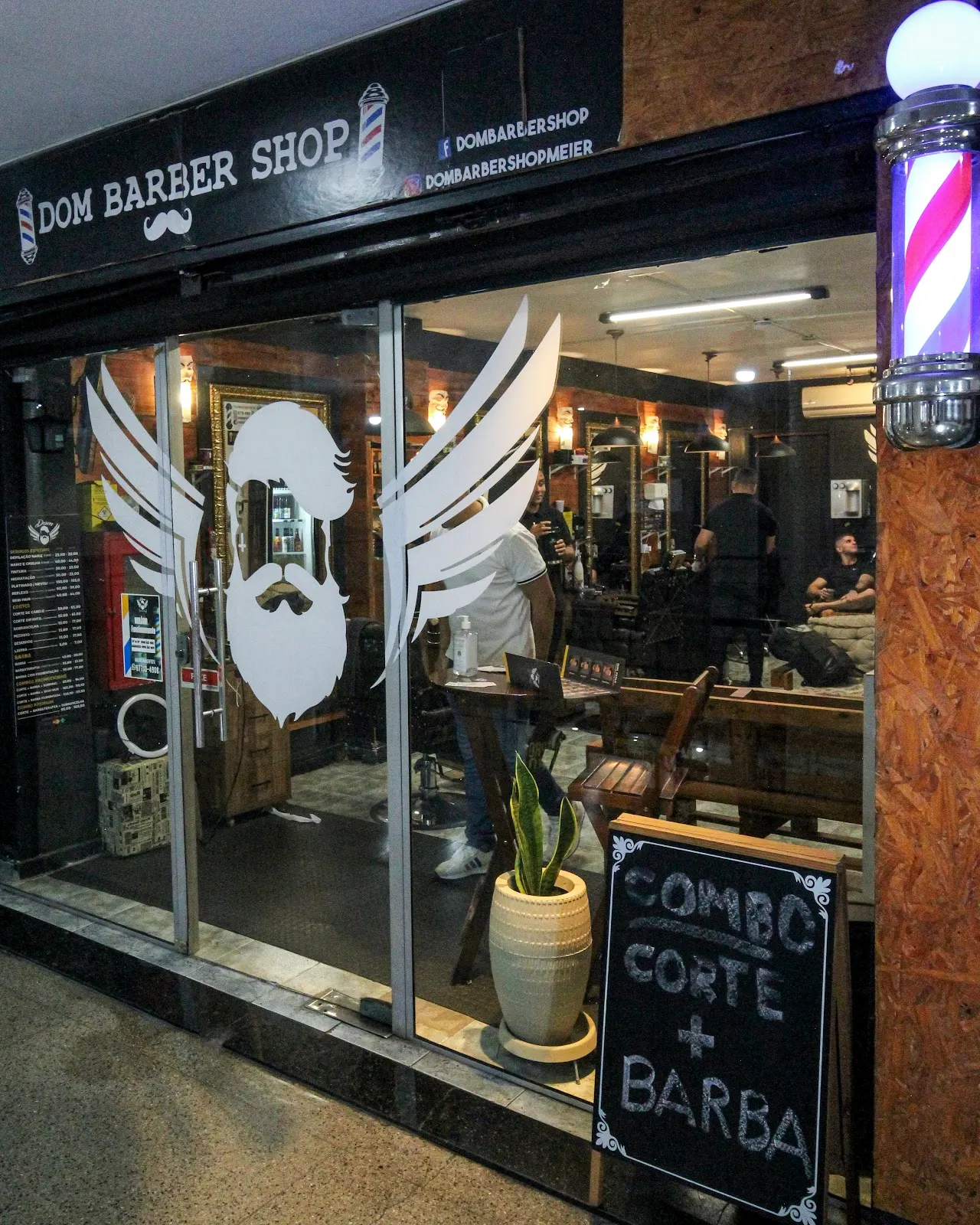 Fachada da Dom Barber Shop à noite, com o logo de asas na vitrine